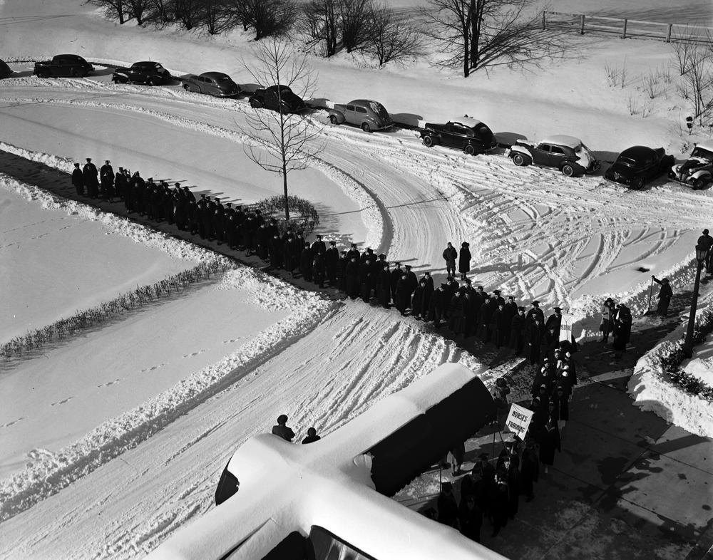  Black and white photograph of cars parked on a snowy street, with a line of people in graduation gowns in the foreground. 