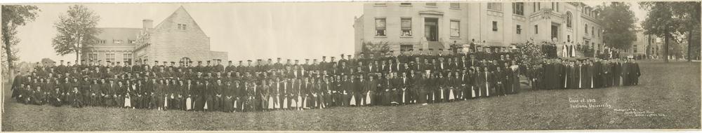  Black and white photograph of students in graduation gowns outside of two campus buildings. 